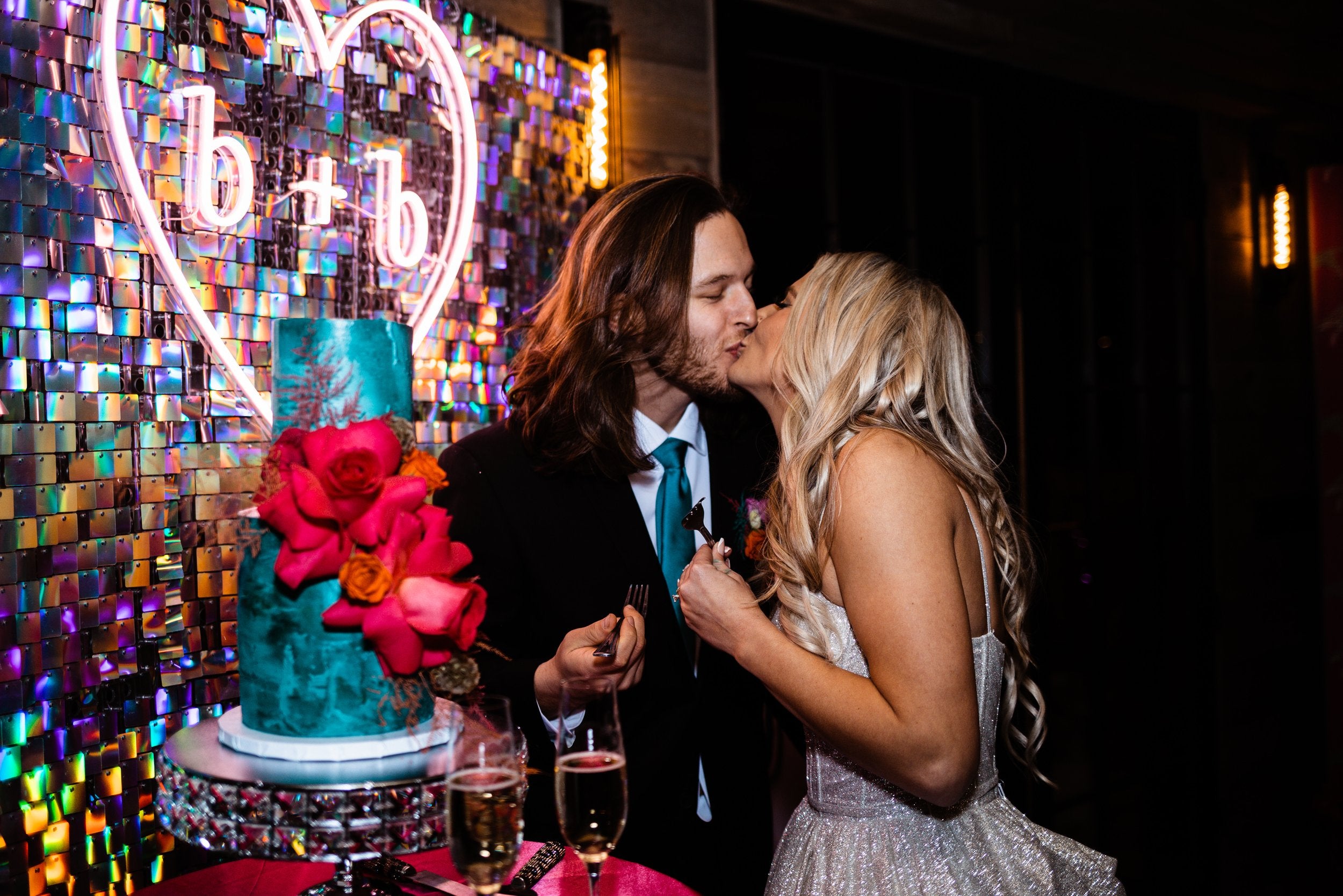 Couple kissing in front of a colorful cake with 'b+b' neon sign in the background