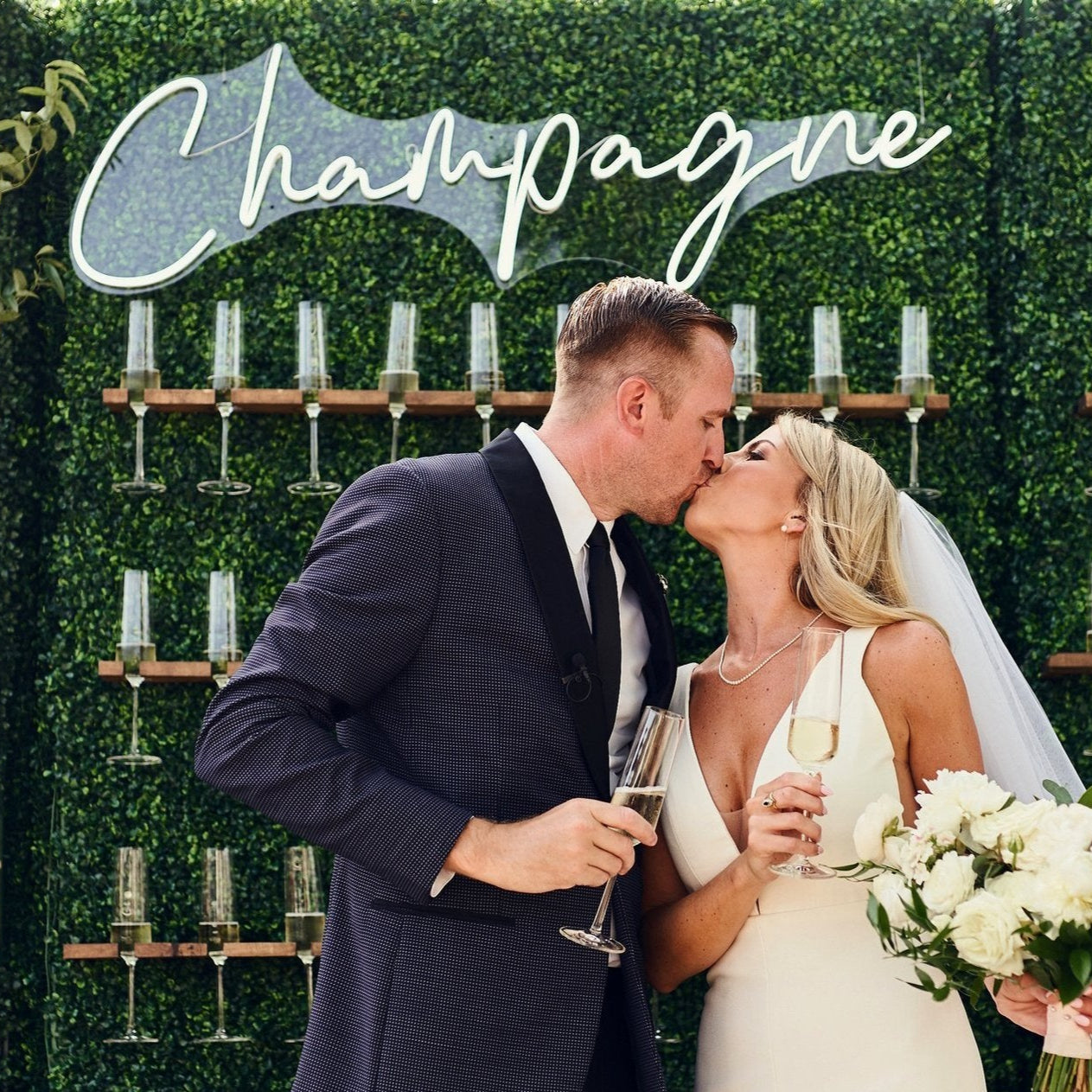 Couple kissing in front of a 'Champagne' sign with champagne bottles on a green wall.
