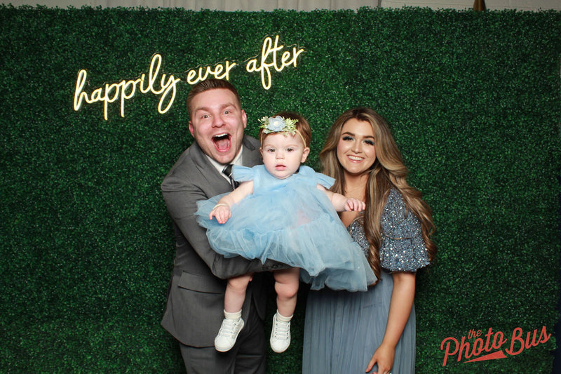 Family of three posing in front of a 'happily ever after' sign with a green backdrop.