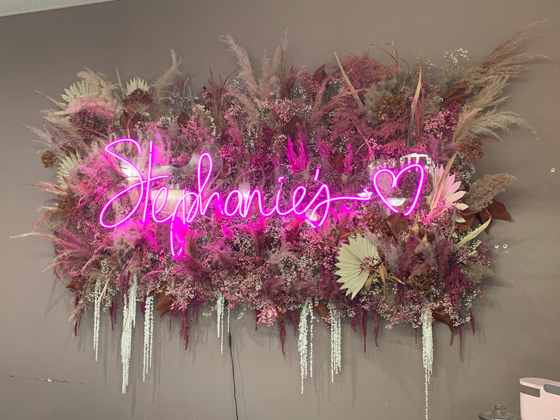 Neon sign with 'Stephanie's' surrounded by decorative flowers and plants on a gray wall.