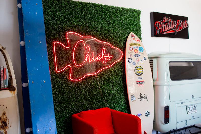 Neon Chiefs sign on a grass-textured wall with a surfboard and van in the background.