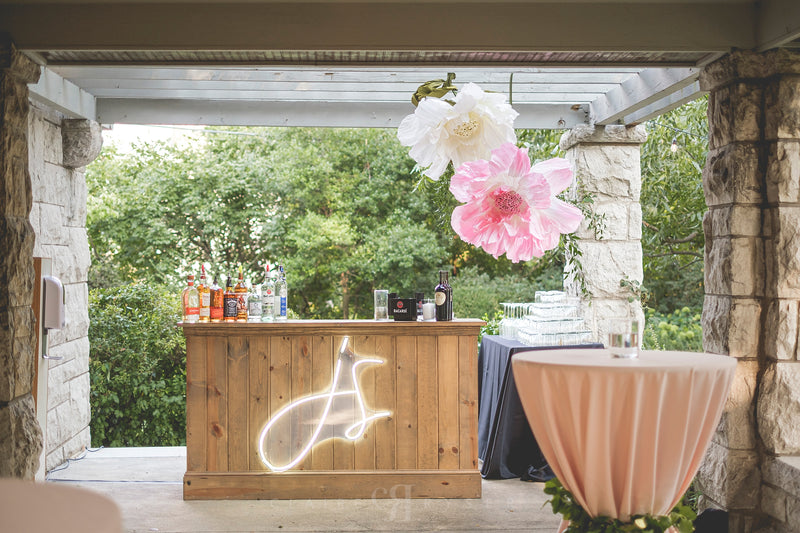 Wooden bar with decorative flowers and bottles, set against a stone wall and greenery.